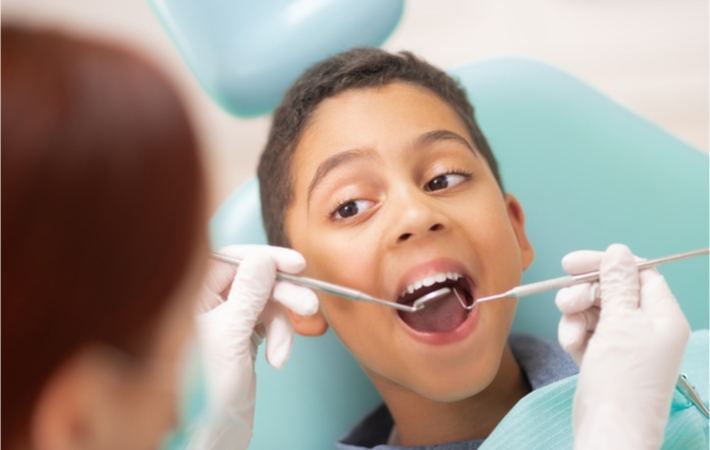 A young boy sitting in a light blue examination chair is looking at the dentist and opening wide as the dentist examines him with a mouth mirror and dental probe.