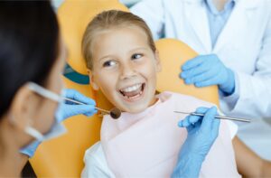 A young smiling girl sitting in an orange examination chair at the dental clinic, facing towards the dentist as she prepares to examine the girl's teeth with a mouth mirror and dental probe.