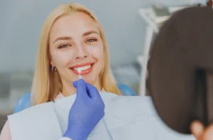Smiling female patient sits in a dentist's chair at the clinic holding up a mirror while the dentist holds up a veneer to her tooth.