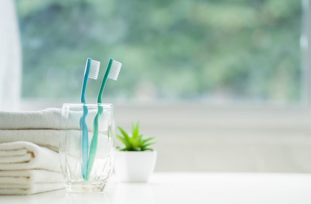 two toothbrushes sitting inside a glass on a white counter top, next to some hand towels and a plant