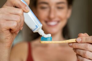 A woman smiling and holding her toothbrush up to the camera while she puts toothpaste onto the brush.