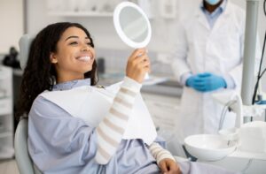 A woman sitting in a dentist's chair admiring her smile in a hand mirror after a dental procedure.