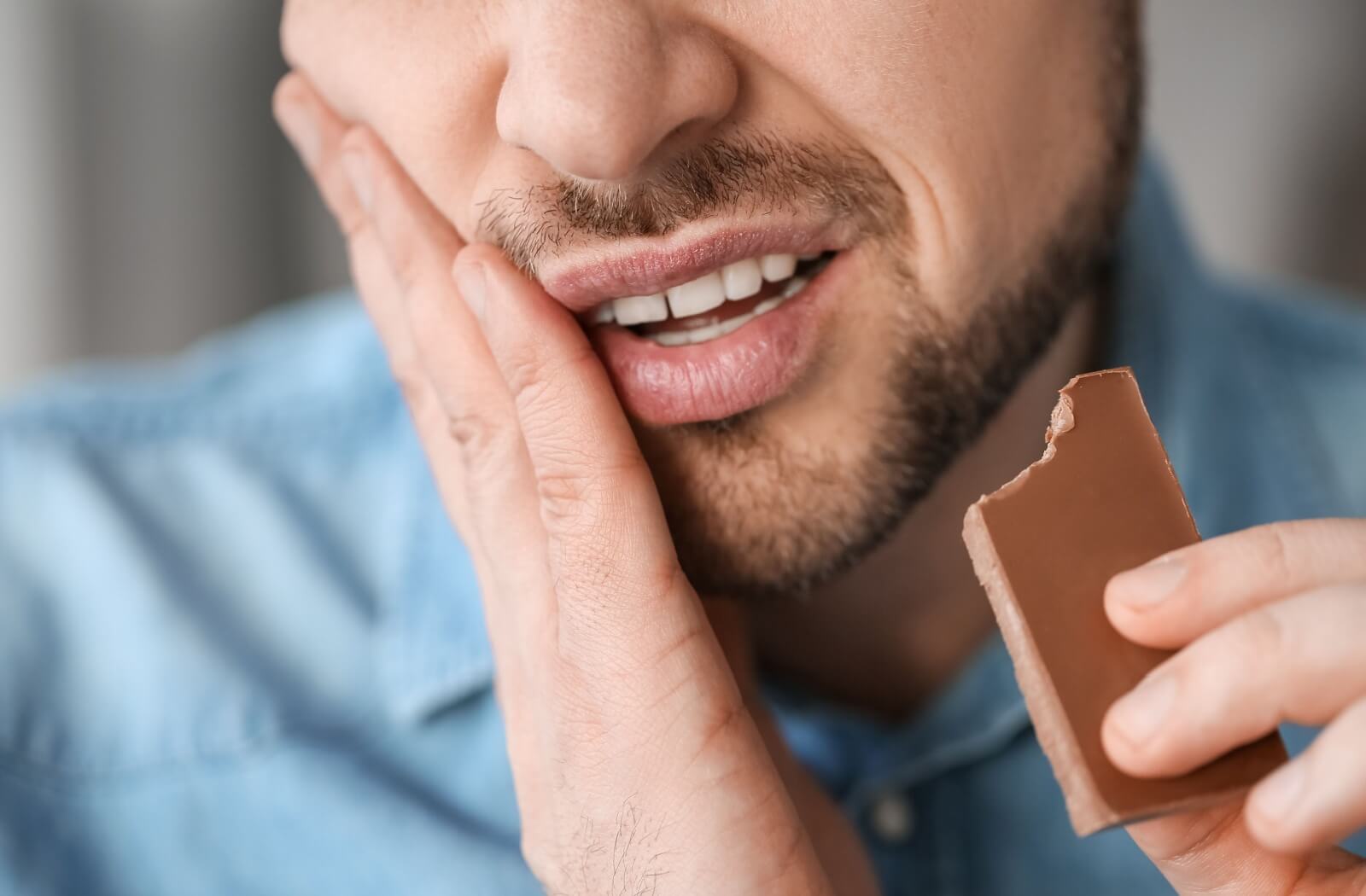 Man holding his jaw in pain while eating chocolate, possibly experiencing tooth sensitivity.