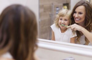 A smiling mother helps her young child brush their teeth while looking into the bathroom mirror.