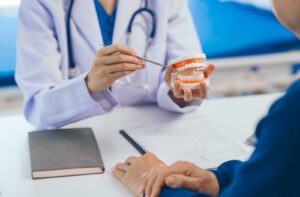 Dental professional holding a tooth model and using a dental tool to explain treatment to a patient seated across the table.