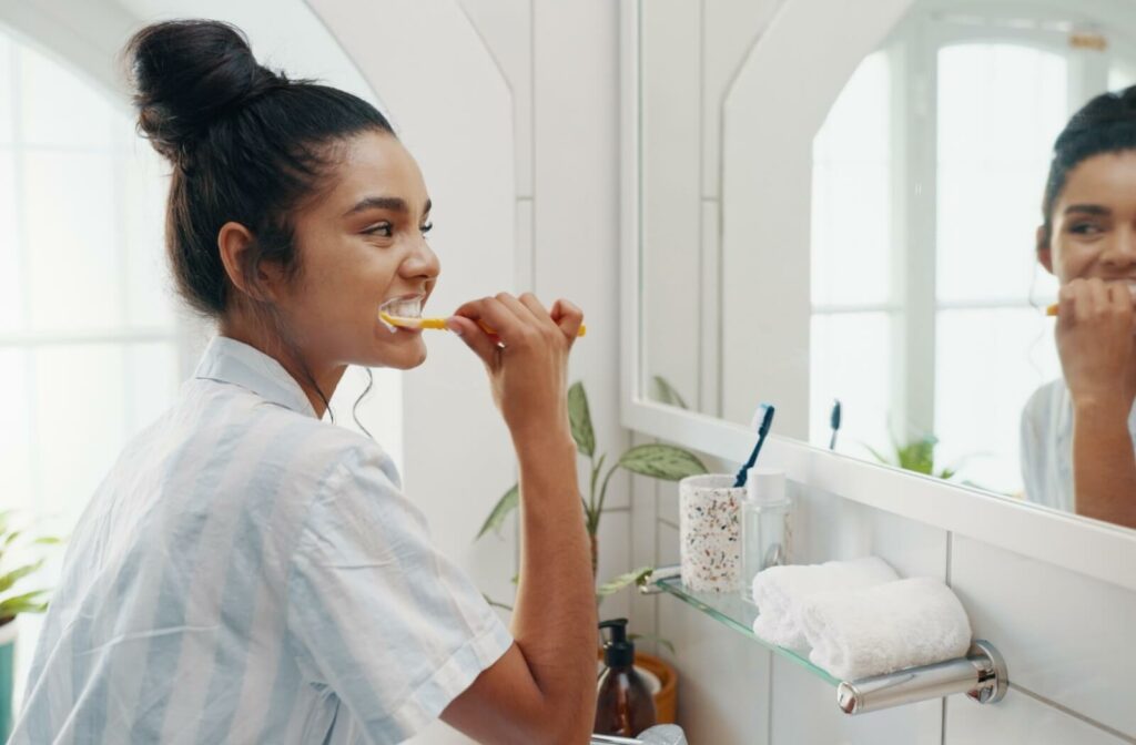 Person brushing their teeth in a bright bathroom while looking into the mirror, with toothbrushes and towels on the counter.