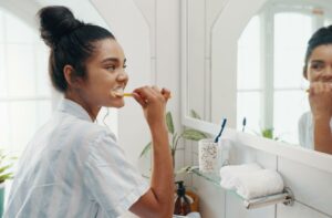 Person brushing their teeth in a bright bathroom while looking into the mirror, with toothbrushes and towels on the counter.