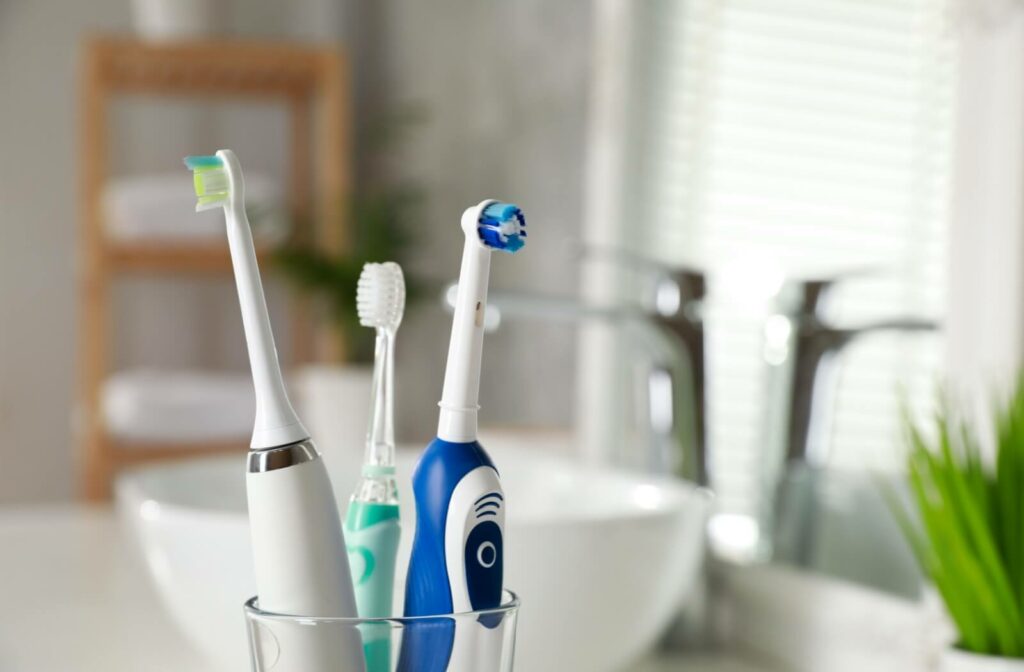 Three toothbrushes, including two electric and one manual, standing in a cup beside a bathroom sink