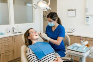 Dentist examines an adult patient's teeth in a dental chair while the patient wears a blue bib.